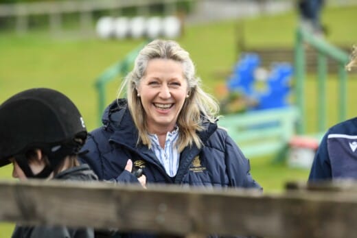 A blonde haired lasy smiling at the camera with horse jumps in the background